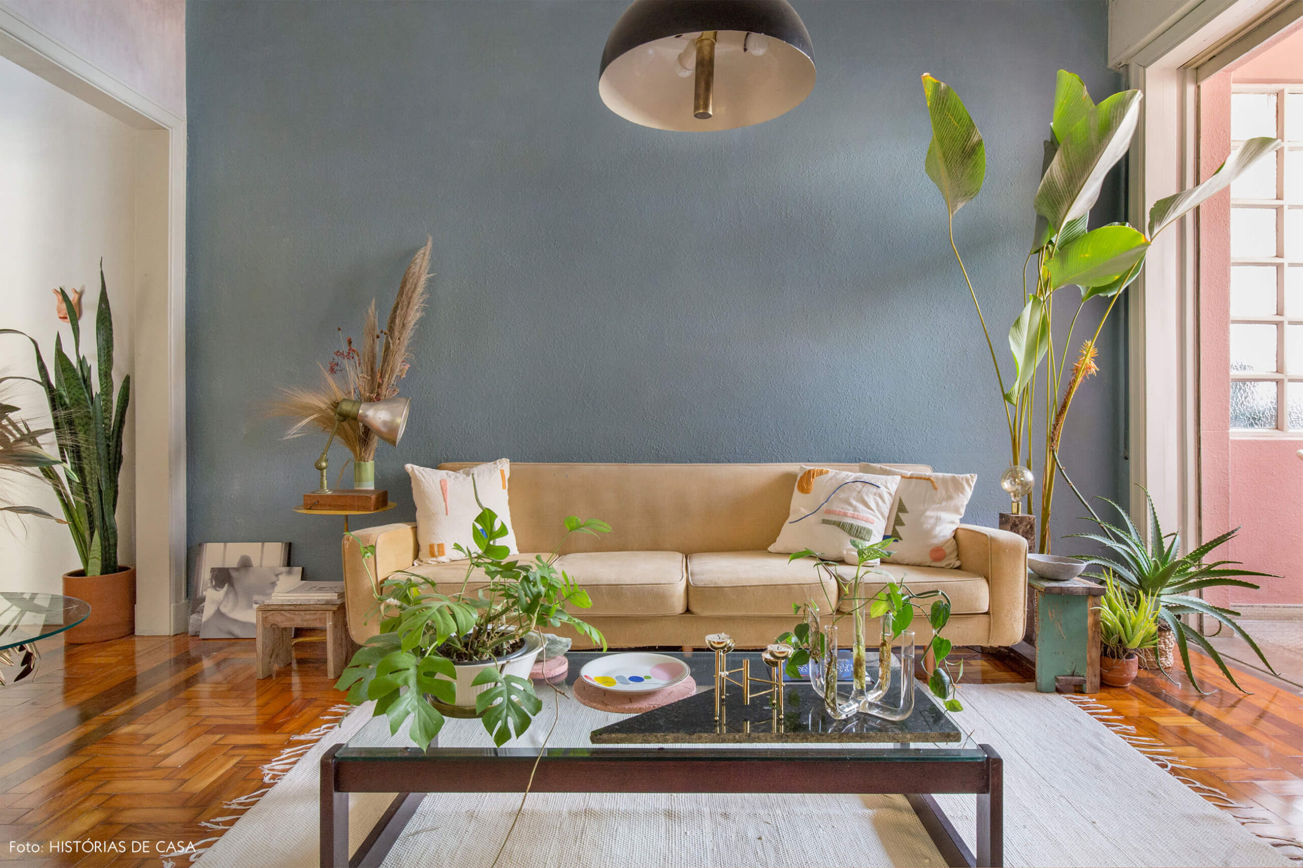 Sala com sofá bege e almofadas bordadas parede azul tapete branco e plantas